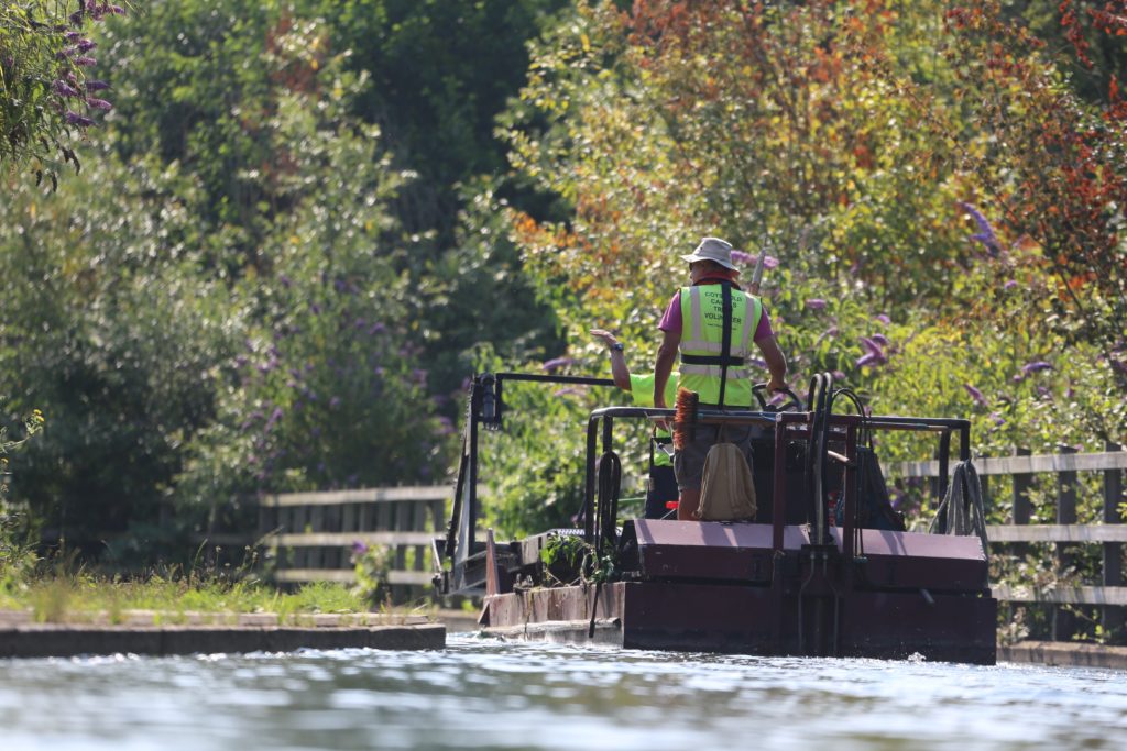 Volunteer in a high-visability jacket on a boat in the canal, clearing debry. Lots of trees surround the canal. 