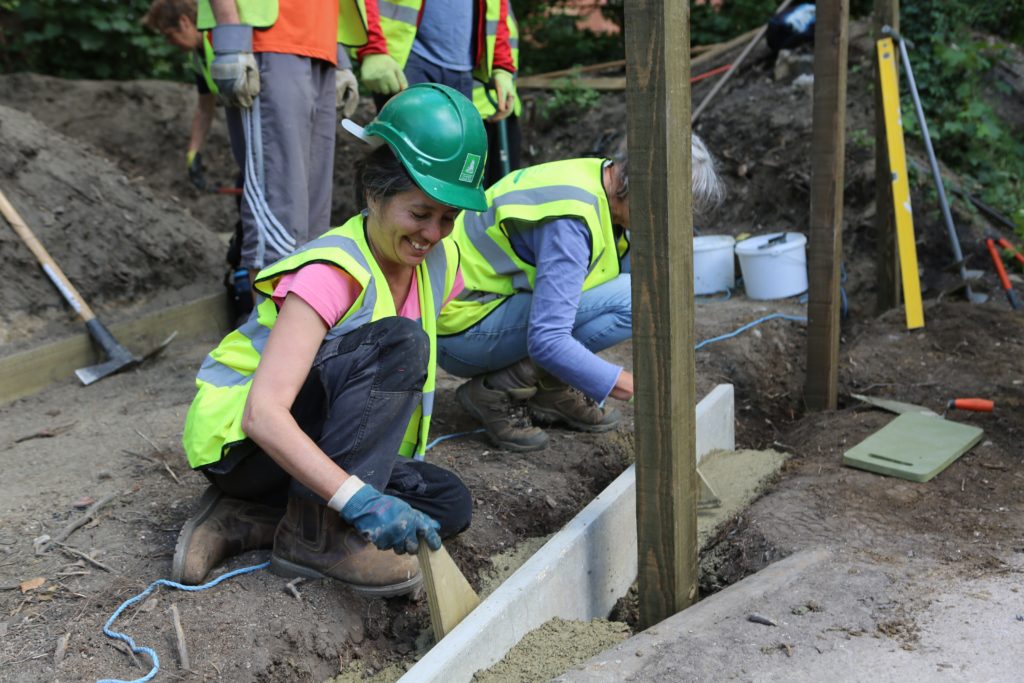Volunteer smiles as she builds the foundation of a pontoon. She wears a hard hat and high-vis jacket. 